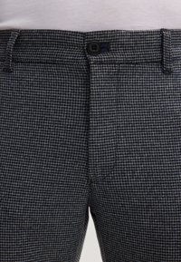 Nahaufnahme von grau-schwarzen Hahnentrittmusterhosen mit Knopfverschluss und Gürtelschlaufen am Bund.