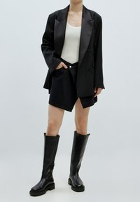 Schwarzer, doppelt geknöpfter Blazer über einem weißen Rippenshirt, kombiniert mit einem schwarzen Jeansrock und knielangen schwarzen Lederstiefeln.