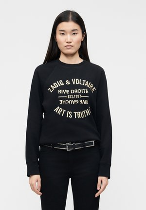Femme portant un sweat-shirt noir avec un texte circulaire beige "Zadig & Voltaire Rive Droite Est.1997 Art is Truth" et une ceinture noire cloutée.
