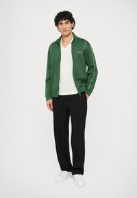 Green zip-up jacket with white piping, worn over a light gray shirt. Paired with black pants and white sneakers.