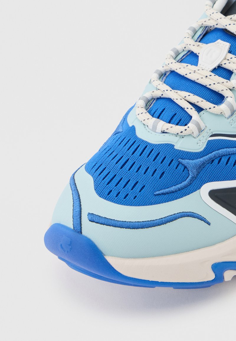 Zapatilla deportiva azul en la parte frontal con sobreposiciones azul claro, cordones gruesos blancos y azules, y detalles de tejido de malla perforada.