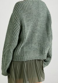 Vihreä neulottu löysä neule, jossa on teksturoitu ribbineulos ja kaapeliyksityiskohtia hihoissa; yhdistetty oliivinvihreään laskostettuun hameeseen.