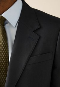 Schwarze Anzugjacke mit strukturiertem Revers, kombiniert mit einem hellblauen Hemd mit Kragen und einer gewebten grünen Krawatte. Verfügt über eine Brusttasche.