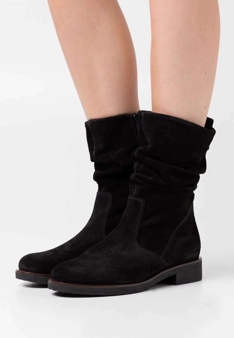 Jambes portant des bottes en daim noir mi-mollet, souples, avec des talons carrés bas et des fermetures éclair latérales sur fond blanc.