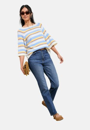 Woman wearing striped pastel sweater, blue jeans, brown flats, sunglasses, and holding a small brown clutch, posing against white background.