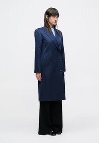 Dvojradový námornícky kabát z hladkej tkaniny, s výrezovými klopami, dlhými rukávmi a bočnými vreckami, doplnený širokými čiernymi nohavicami.