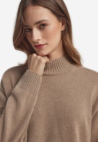 Beżowy sweter z wysokim kołnierzem, z prążkowymi ściągaczami na mankietach i kołnierzu, wykonany z miękkiego, teksturowanego dzianiny. Zbliżenie na twarz i dłonie modela.