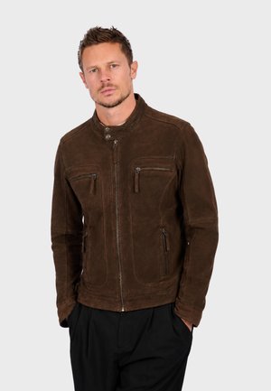 Homme portant une veste zippée en daim marron avec des poches zippées sur la poitrine et les côtés, debout les mains dans les poches d’un pantalon noir.