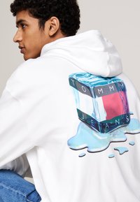 Felpa bianca con cappuccio caratterizzata da una grafica di un cubetto di ghiaccio con la scritta "TOMMY JEANS" sopra, con un effetto di vernice blu che si scioglie sotto il cubo.