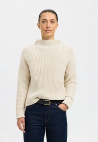 Pull beige en tricot avec un col haut côtelé et des manches longues, associé à un jean en denim foncé et une ceinture noire avec une boucle dorée.