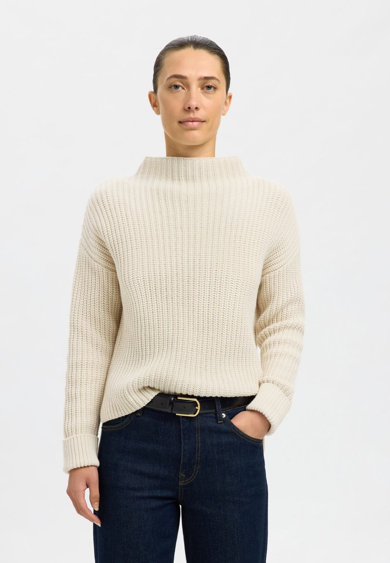 Pull beige en tricot avec un col haut côtelé et des manches longues, associé à un jean en denim foncé et une ceinture noire avec une boucle dorée.