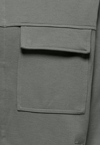 Gray fabric square pocket with flap and visible stitching on textured garment surface.