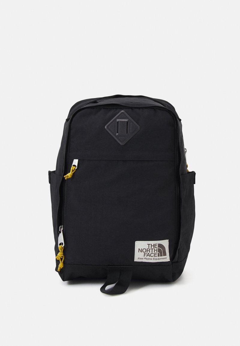 Sac à dos rectangulaire noir avec des fermetures éclair à l'avant, des tirettes jaunes, des poches latérales et un patch logo North Face blanc en bas à droite.
