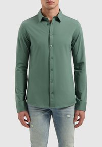 Langsleeve overhemd in groene stof, met een kraag, teal knopen en een gladde textuur. Gecombineerd met lichtblauwe distressed jeans.