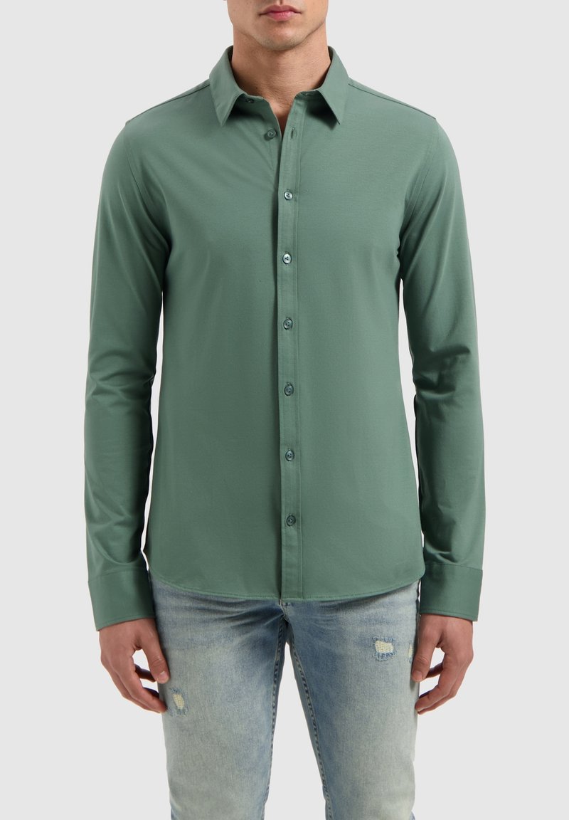 Langsleeve overhemd in groene stof, met een kraag, teal knopen en een gladde textuur. Gecombineerd met lichtblauwe distressed jeans.
