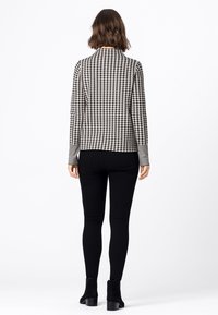 Woman with short wavy hair wearing a black and white houndstooth jacket, black skinny pants, and black ankle boots, standing facing away.