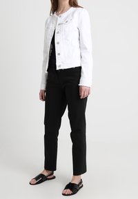 Giacca di denim bianco con chiusura a bottoni, due tasche sul petto e orlo sfilacciato. Abbinata a pantaloni neri e sandali neri.