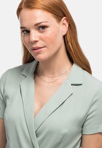 Hellgrüne Wickelbluse mit kurzen Ärmeln, strukturiertem Stoff; geschichtete goldene Halsketten mit kleinen weißen Perlen betonen den Ausschnitt.