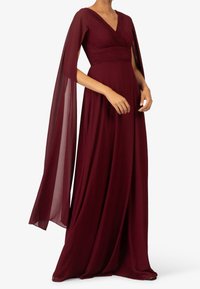 Nainen, joka on pukeutunut lattian pituiseen, syvän punaiseen mekkoon, jonka runko on laskostettu ja jossa on virtaavat läpikuultavat hihansuut valkoista taustaa vasten.