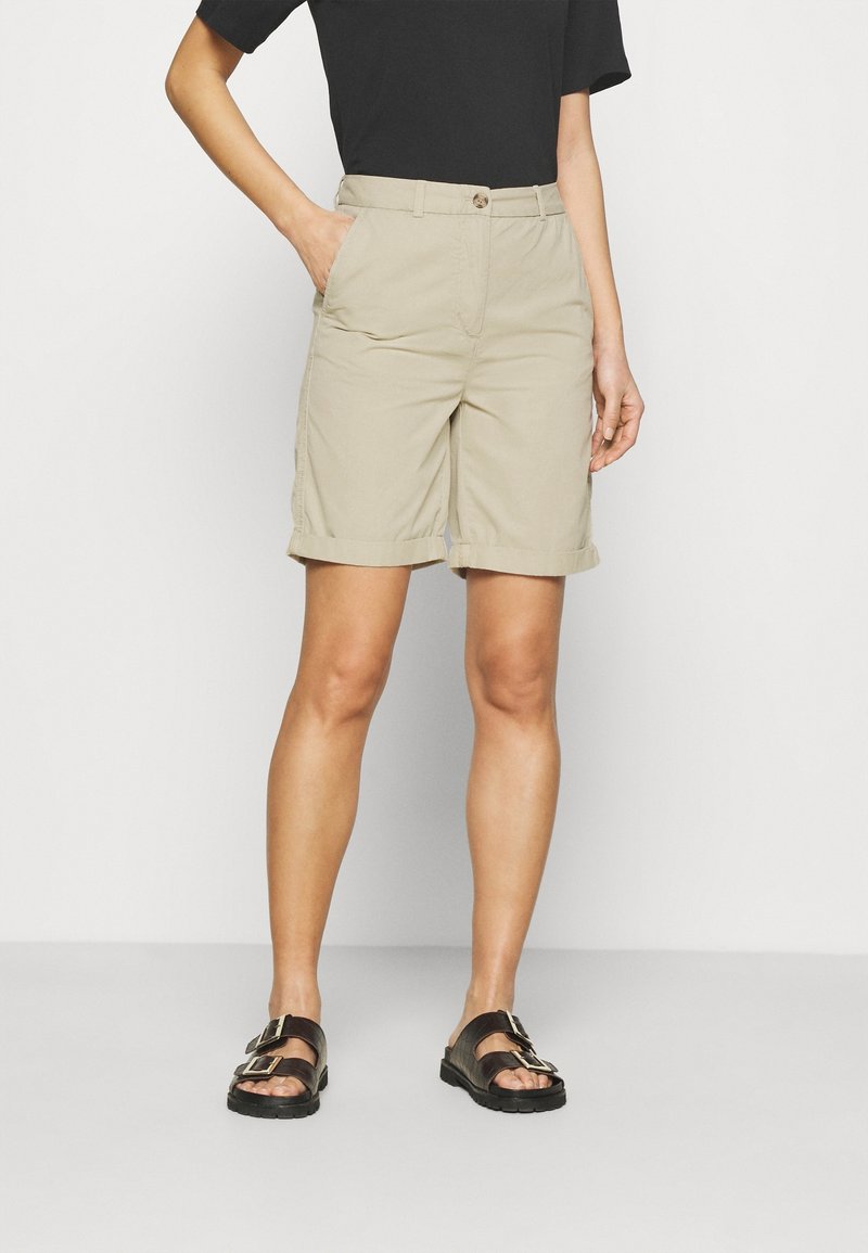 Pantaloni corti beige in cotone, con chiusura a bottone, passanti per cintura e tasche laterali. Indossati con sandali neri e una maglietta nera.