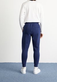 Marina joggerbyxor med en avsmalnande design, elastisk midja och bakficka; kombineras med en vit långärmad skjorta och vita sneakers.