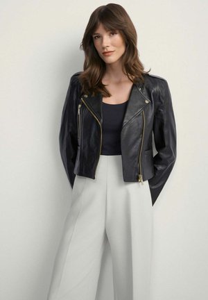 Kobieta o brązowych włosach, ubrana w czarną skórzaną kurtkę, czarny top i białe, szerokie spodnie z wysokim stanem, stojąca na jednolitym tle.