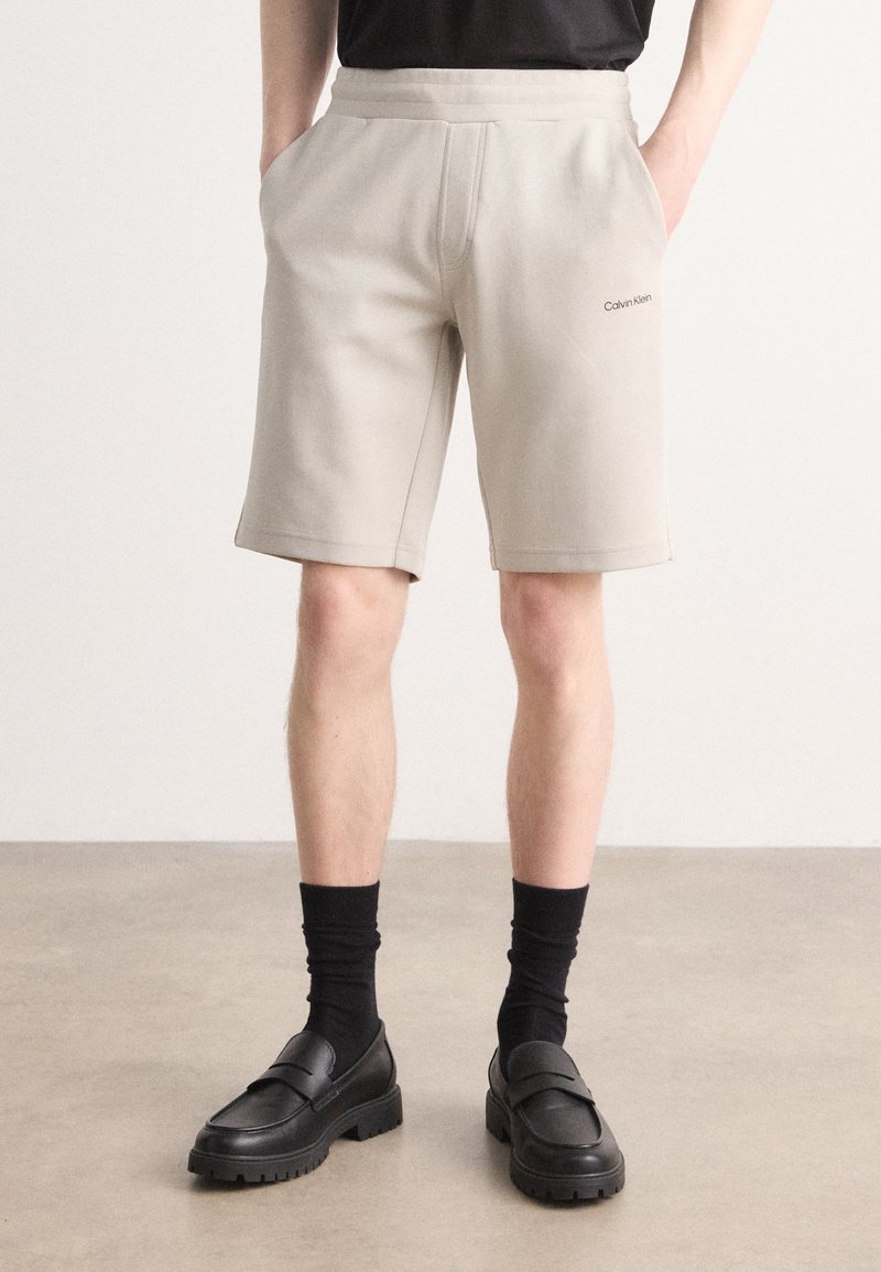 Shorts en coton beige mélangé avec une taille élastique, des poches latérales et un logo discret. Stylisés avec des mocassins noirs et des chaussettes.