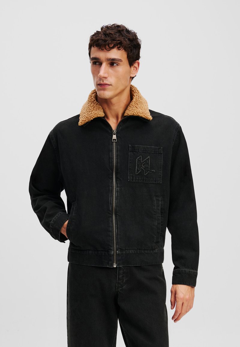 Chaqueta de mezclilla negra con un cuello de piel sintética en tono beige, cierre de cremallera y bolsillo frontal con logotipo bordado. Tejido texturizado con un corte relajado.