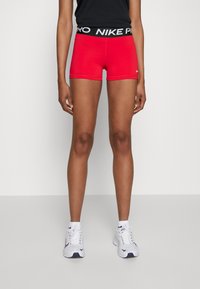 Shorts Nike Pro rouges avec une ceinture contrastante noire portant la mention "NIKE PRO" en lettres en gras. Tissu lisse et extensible, conçu pour le confort.