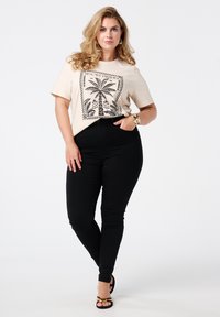 Beige t-shirt met zwart palmbomenprint, gecombineerd met hoge zwarte skinny jeans en zwarte sandalen met gouden accenten.