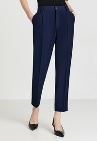 Marineblaue Anzughosen mit Falten, Knopfverschluss, Seitentaschen und schmal zulaufenden Beinen. Kombiniert mit schwarzen hochhackigen Schuhen. Glatte Stoffoberfläche.
