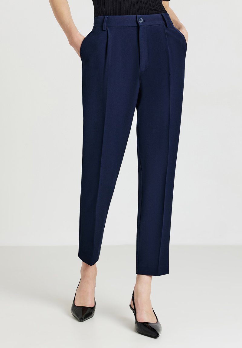 Pantaloni da cerimonia blu con pieghe, chiusura a bottone, tasche laterali e gambe affusolate. Abbinati a scarpe nere con tacco alto. Tessuto dalla texture liscia.