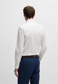 Camicia bianca a maniche lunghe con colletto classico, tessuto liscio e polsini con bottoni, abbinata a pantaloni blu scuro con tasche posteriori.