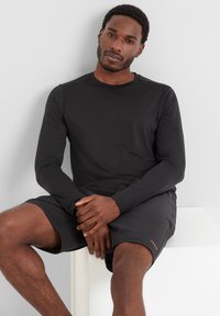 Black long-sleeve shirt made of smooth fabric, paired with black shorts featuring a small red logo on the hem. Model seated with hands crossed.