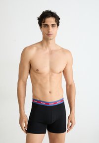 Bóxer de algodón negro con una cinturilla elástica de contraste azul y rojo, que presenta el logotipo de la marca "HUGO" repetido alrededor de la banda.