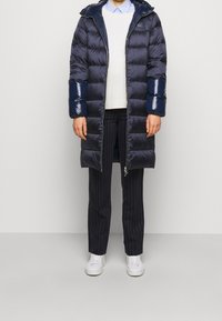 Marinblå quiltad pufferjacka med glansiga blå detaljer, huva och dragkedja fram. Bärs över en ljus tröja och randiga byxor. Vita sneakers.