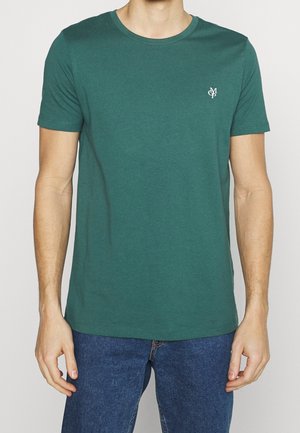 Dorosła osoba ubrana w zieloną, gładką koszulkę z krótkim rękawem z małym białym logo "MSP" na lewej piersi, w połączeniu z niebieskimi dżinsami.