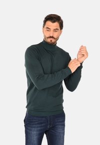 Maglione dolcevita verde scuro, materiale morbido a maglia, design aderente, collo e polsini a coste, abbinato a jeans di denim.