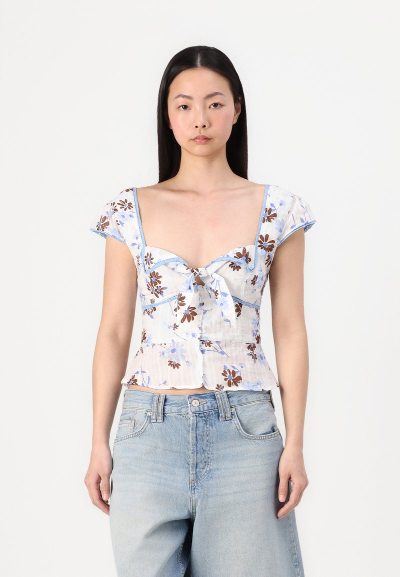 Blusa a stampa floreale con maniche corte a sbuffo, dettaglio con fiocco frontale e accenti blu chiaro. Il materiale sembra leggero e presenta una finitura testurizzata.