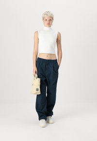 Top bianco senza maniche, pantaloni blu navy larghi e una piccola borsa beige con una toppa. Sneakers bianche completano l'outfit. Design minimalista.