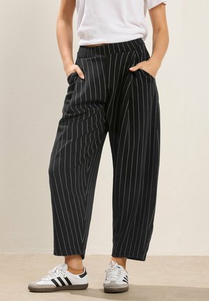 Personne portant un pantalon noir à jambes larges avec des fines rayures blanches et des baskets blanches, debout les mains dans les poches sur un fond neutre.