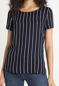 Frau in einem marineblauen Kurzarmhemd mit vertikalen weißen Streifen, kombiniert mit blauen Jeans, steht vor einem schlichten Hintergrund.