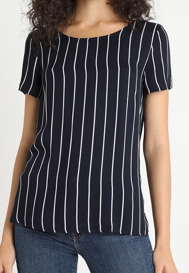 Mujer vestida con una camisa de manga corta azul marino con rayas verticales blancas, combinada con jeans azules, de pie frente a un fondo liso.