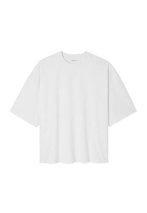 Jeune homme aux cheveux courts et bouclés porte un t-shirt blanc ample et un short bleu clair, debout devant un fond clair uni.