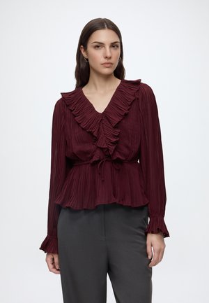 Femme aux longs cheveux foncés portant un chemisier bordeaux à volants avec des plis et un pantalon gris foncé, debout devant un fond clair uni.