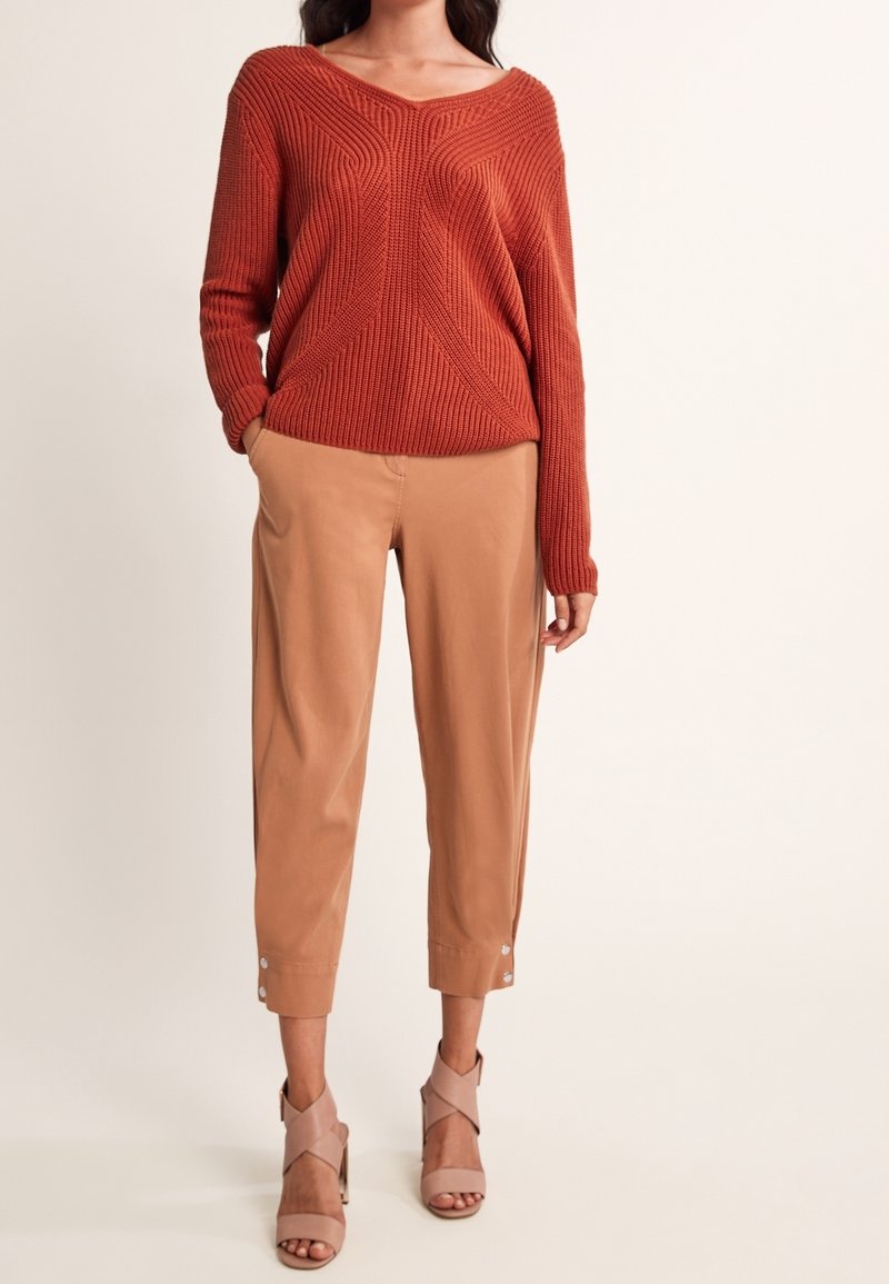 Femme portant un pull en tricot couleur rouille, un pantalon court beige avec des boutons aux poignets, et des sandales à talons hauts beige à bout ouvert, se tenant devant un fond uni.