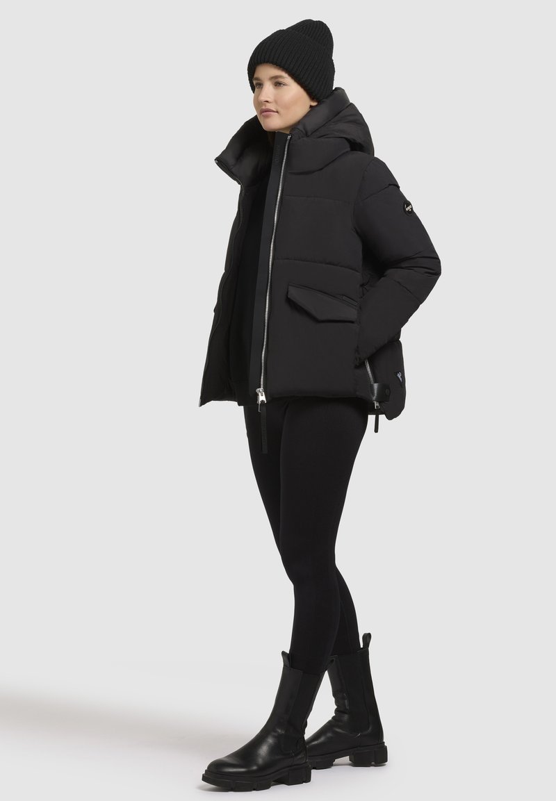 Schwarze Pufferjacke mit hohem Kragen, Frontreißverschluss und zwei Seitentaschen. Kombiniert mit einer schwarzen Strickmütze und eng anliegenden schwarzen Leggings.