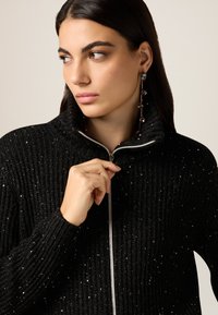 Schwarzer Strickpullover mit Reißverschluss und hohem Kragen, mit einer gerippten Struktur und silbernen Pünktchen. Das Modell trägt Ohrringe mit Perlen.