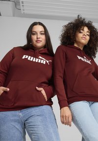 Felpe maroon con grande logo bianco "PUMA", dotate di tasche a marsupio e cappucci con cordino, abbinate a jeans blu.