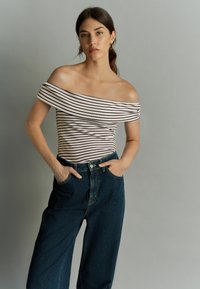 Off-the-shoulder, geribbelde top in lichtroze met donkere strepen, gecombineerd met hooghangende donkere denim broek met voorzakken.